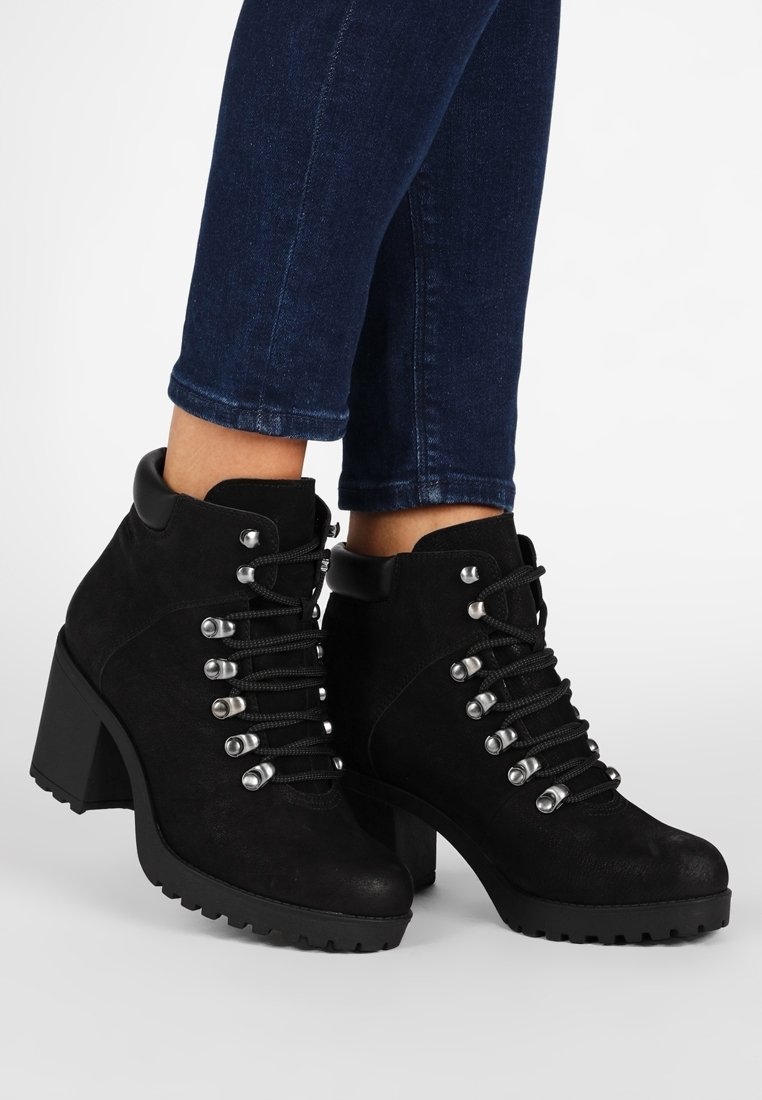 Zwarte suède enkellaarsjes met een stevige hak, voorzien van metalen oogjes, zwarte veters en een textuurrijke zool voor extra grip.