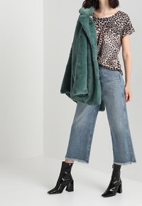 Abrigo de pelaje sintético verde, camiseta de estampado animal, jeans cortos de pierna ancha de denim claro y botines negros de charol con tacones cuadrados.