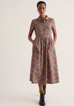 Femme debout face à l'avant, mains dans les poches, portant une robe midi à fleurs à manches courtes, des bottes noires et des chaussettes jaunes sur un fond uni.