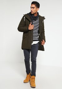 Oliwkowa parka z kapturem, zapinana na zamek i guziki, warstwowa na czarna i białą koszulę w paski, zestawiona z szarymi rurkami i żółtymi butami.