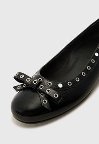 Black leather ballet flat with a rounded toe, featuring a bow and silver eyelet accents around the opening. Textured finish, cushioned interior.