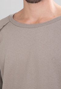 Sudadera de algodón color beige claro con cuello redondo de ribete y una ligera textura; presenta un pequeño logo bordado en el pecho.