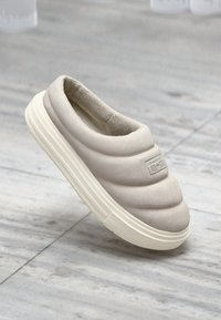 Lys beige slip-on sko med polstert overdel, teksturert overflate og kremfarget gummisåle. Akkompagnert av en liten logo på overpanelet.