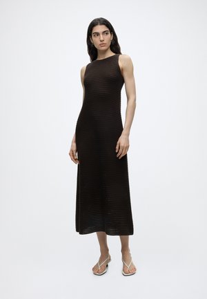 Kobieta z długimi ciemnymi włosami, ubrana w czarną dzianinową sukienkę midi bez rękawów i białe płaskie sandały na paski, stojąca na jednolitym tle.