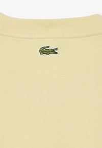 T-shirt in cotone giallo con scollatura rotonda e un piccolo logo di coccodrillo verde ricamato sul retro. Texture liscia, design casual.