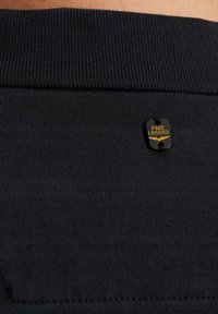 Tela negra con textura acanalada y una pequeña etiqueta metálica con "PME Legend" en dorado, unida en la zona del cuello.