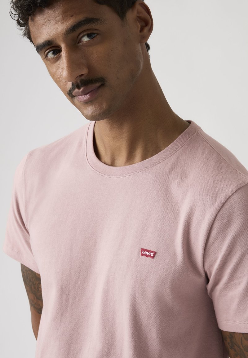Homme avec moustache et cheveux bouclés portant un t-shirt Levi’s rose clair, face à l’avant avec une expression neutre sur un fond uni.