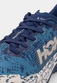 Zilas skriešanas apavi ar teksturētu auduma augšdaļu, kontrastējošām zilām auklām un baltu zoli. Sānos ir redzams logo un modeļa nosaukums "SPEEDGOAT 6".