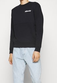 Svart sweatshirt med rund halsringning, långa ärmar och en texturerad panel. Har en vit "NikeAir"-logotyp på bröstet. Tillsammans med ljusblå jeans.