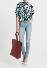 Donna che indossa una blusa floreale e jeans skinny azzurri chiari, mentre tiene una grande borsa a tracolla rossa, abbinata a scarpe argentate con tacco a punta.