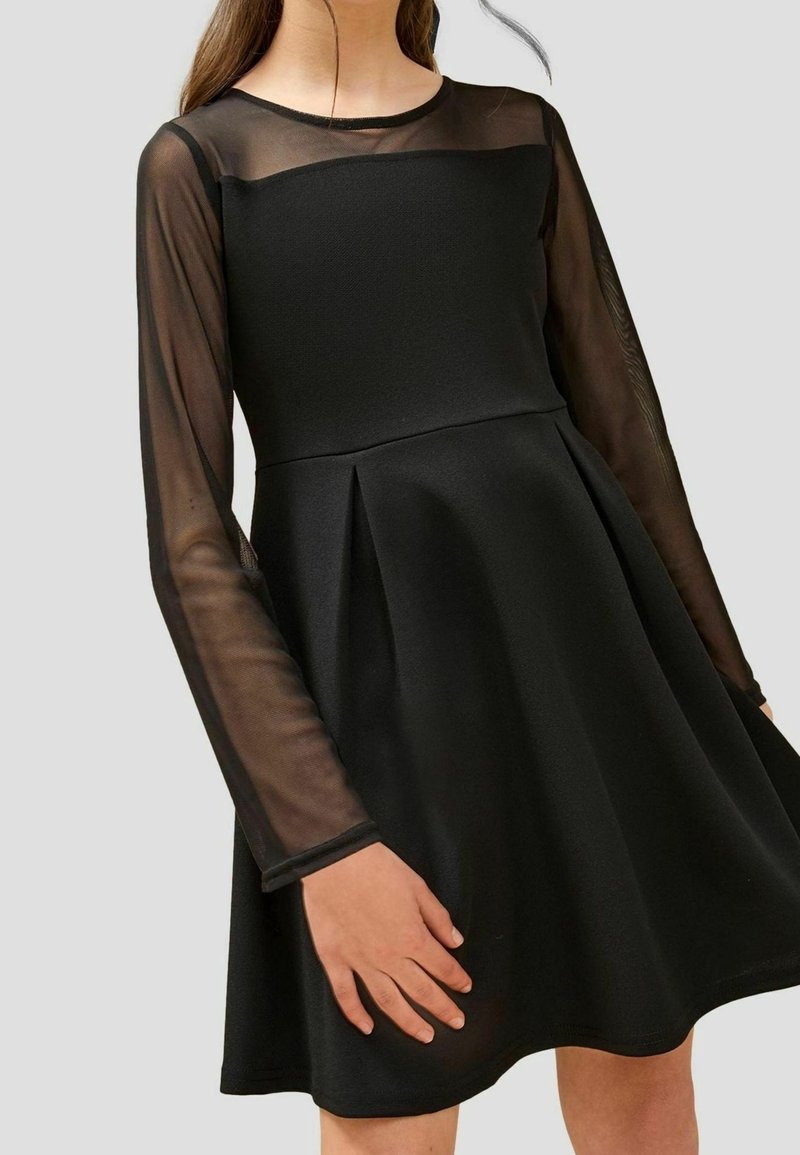 Robe noire avec corsage ajusté, jupe évasée, manches longues transparentes et panneau supérieur de poitrine sur une personne.