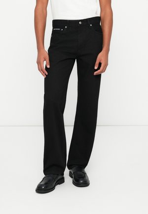 Mężczyzna ubrany w czarne jeansy o prostych nogawkach, czarne skórzane mokasyny oraz białą koszulę z krótkim rękawem, stojący na białym tle.