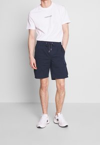 Shorts cargo bleu marine avec taille à cordon, dotés de poches latérales et d'une coupe décontractée, assortis à un t-shirt en coton blanc avec logo.