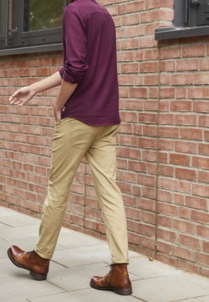Stivali in pelle marrone, pantaloni beige e una camicia a maniche lunghe viola scuro con maniche arrotolate. L'individuo cammina su una superficie pavimentata accanto a un muro di mattoni.