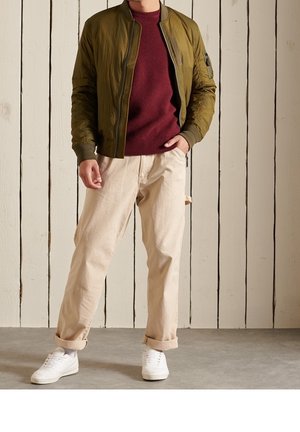 Veste bomber vert olive portée sur un pull texturé bordeaux, associée à un pantalon crème fuselé et des baskets blanches.