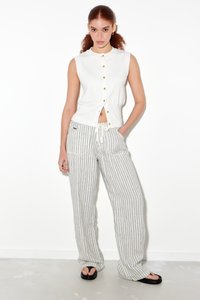 Hvit ermeløs topp med gullknapper, kombinert med lyse stripete, vide bukser. Fottøy er svarte sandaler. Minimalistisk design, uformell bekledning.