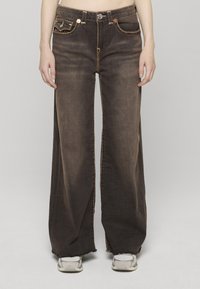 Džíny s širokými nohavicemi v tmavě modrém denimovém provedení s kontrastním žlutým prošíváním, s předními kapsami a třepenými lemy. Stylizováno s lehkými teniskami.