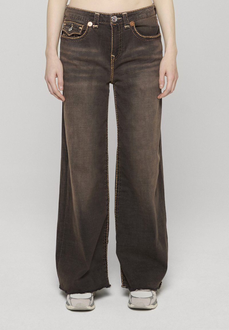 Džíny s širokými nohavicemi v tmavě modrém denimovém provedení s kontrastním žlutým prošíváním, s předními kapsami a třepenými lemy. Stylizováno s lehkými teniskami.