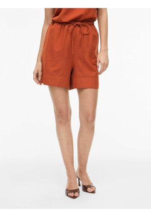 Vrouw draagt roestkleurige linnen shorts met trekkoord in de taille en bruine open-toe sandalen met hak, staand tegen een witte achtergrond.