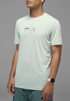 Homme portant un T-shirt Millet gris clair à manches courtes avec un texte noir et un pantalon bleu sarcelle, debout devant un fond gris.