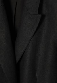 Černý blazer s texturovanou, tkanou látkou. Obsahuje špičaté klopy, přiléhavý střih a dvě přední kapsy. Klasický design a barva.