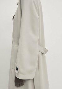 Jasnoszary płaszcz o uporządkowanej konstrukcji, z długimi rękawami, detalem w postaci guzika na nadgarstku oraz akcentem w postaci związanej w talii. Gładka tekstura materiału.