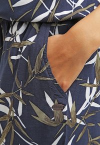 Tissu bleu marine avec un motif feuillu vert et blanc, doté d'une poche latérale et d'un accent métallique avec le nom de la marque « Denim ».