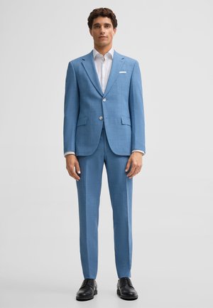 Mężczyzna stojący, ubrany w szyty na miarę jasnoniebieski garnitur z białą koszulą i czarnymi eleganckimi butami na jednolitym jasnym tle.