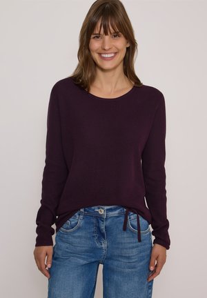 Jeune femme souriante, vêtue d'un pull en maille violet foncé et d'un jean bleu, debout devant un fond clair uni.