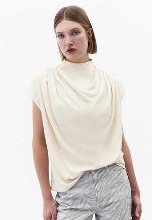 Kobieta o prostych jasnobrązowych włosach nosi kremowy, drapowany top bez rękawów i jasnoszare spodnie w print zebry, pozując na białym tle.