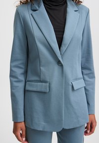 Lichtblauwe op maat gemaakte blazer met twee voorkruiszakken en een enkele knoopsluiting, gemaakt van een gestructureerde stof. Draagt over een zwarte coltrui.