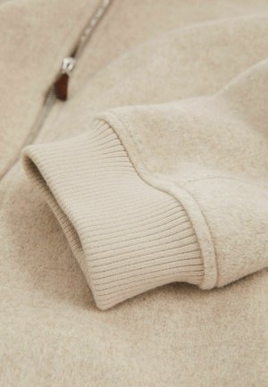 Manșeta puloverului din fleece bej, cu textură tricotată în dungi, având o suprafață netedă și pufoasă, cu un fermoar metalic pe marginea din față.