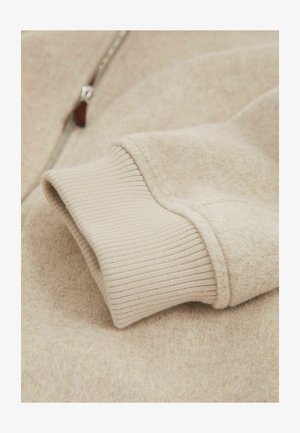 Bežs vilnas džemperis ar ribu adījumu stāvu, kuram ir gluda, pūkaina virsma un metāla rāvējslēdzis gar priekšējo malu.