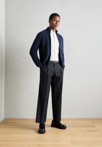 Cardigan a zip con coste in navy, camicia bianca con bottoni, pantaloni larghi neri e scarpe nere. Tessuti lisci e palette di colori neutri.