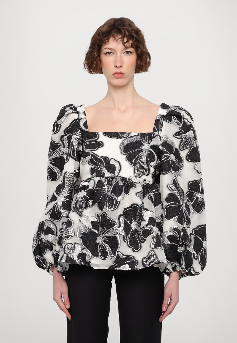 Blusa floral en blanco y negro con mangas abullonadas llamativas, escote cuadrado, tejido texturizado y patrones de flores bordados. Pantalones ajustados negros.