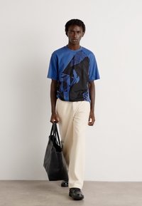 Zila un melna grafiskā T-kreklā, brīvs piegriezums, kombinēts ar krēmkrāsas platām biksēm un melnu somu. Apavi ir melni ar teksturētu dizainu.