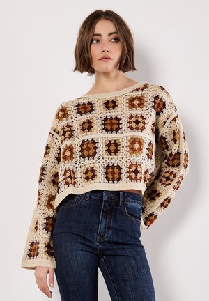 Crochet sweater in cream, brown, and rust with a square pattern, wide sleeves, and a cropped design, paired with dark blue jeans.