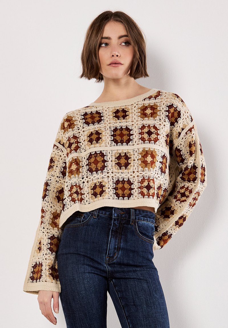 Crochet sweater in cream, brown, and rust with a square pattern, wide sleeves, and a cropped design, paired with dark blue jeans.