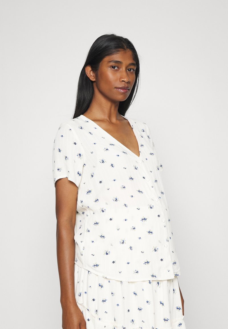 Blusa branca de manga curta com decote em V, apresentando um padrão floral em azul e verde. Tecido leve com um corte descontraído e textura suave.