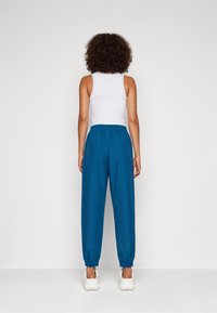 Modré joggingové kalhoty s elastickým pasem a manžetami, vyrobené z měkkého materiálu. Kombinované s bílým tílkem. Pohled zezadu.