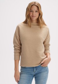 Beige textuur trui met een hoge hals en een relaxed fit, gecombineerd met lichtblauwe jeans, gepresenteerd tegen een effen witte achtergrond.
