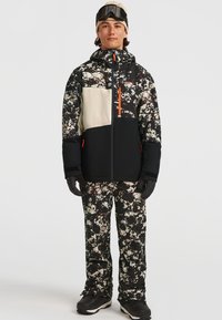 Ski-jack en broekset in zwart, wit en meerkleurig patroon. De jas heeft een grote voorkantzak en oranje accenten. Geïsoleerde stof.