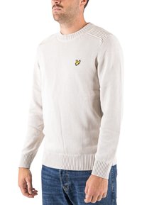 Pull en tricot beige clair avec poignets et ourlet côtelés, présentant un petit logo jaune sur la poitrine. Encolure ronde standard et coupe décontractée.
