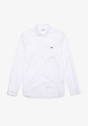 Camisa blanca de manga larga, con botones, confeccionada en tela de algodón. Presenta un cuello extendido, cierre frontal con botones y un pequeño logo verde en el lado izquierdo del pecho.