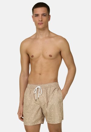 Joven con bañador beige con estampado de paisley y cordón blanco, de pie con una mano en el bolsillo sobre un fondo claro.