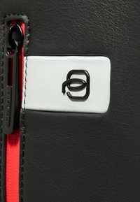 Matériau noir texturé avec un panneau blanc contrastant, arborant un logo noir et une fermeture éclair rouge. Coutures soignées le long des bords.