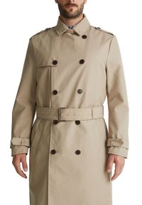 Trench-coat beige à double boutonnage, boutons marron, ceinture à la taille, épaulettes et brides aux poignets, fabriqué en tissu lisse.
