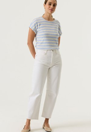 Jeune femme debout, mains derrière le dos, portant un haut à manches courtes rayé bleu et beige, un jean blanc taille haute et des ballerines beiges.