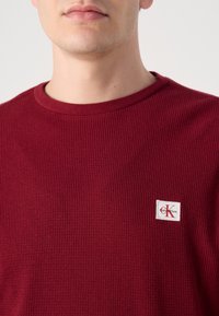 Bordeaux geribbelde gebreide langemouwen shirt met een ronde halslijn en een klein wit Calvin Klein logo label op de borst.
