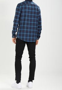 Koszula w kratę w niebiesko-czarnym kolorze, wykonana z tkaniny tkanej; dopasowany krój z zapięciem na guziki przy mankietach. Czarne spodnie slim-fit i białe sneakersy.